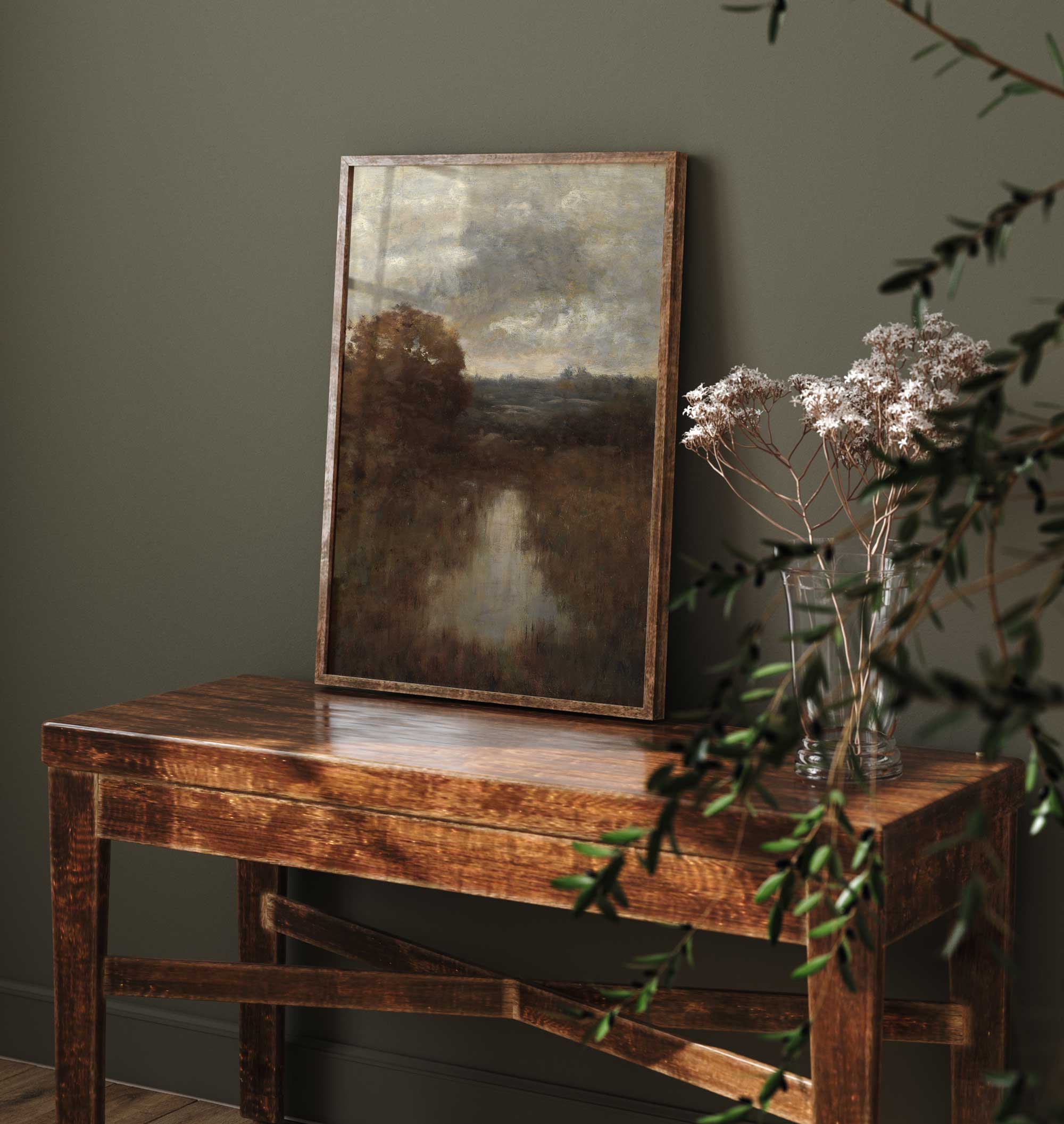 Framed Rustic landscape print on a wooden table with a vase of flowers against a dark green wall.