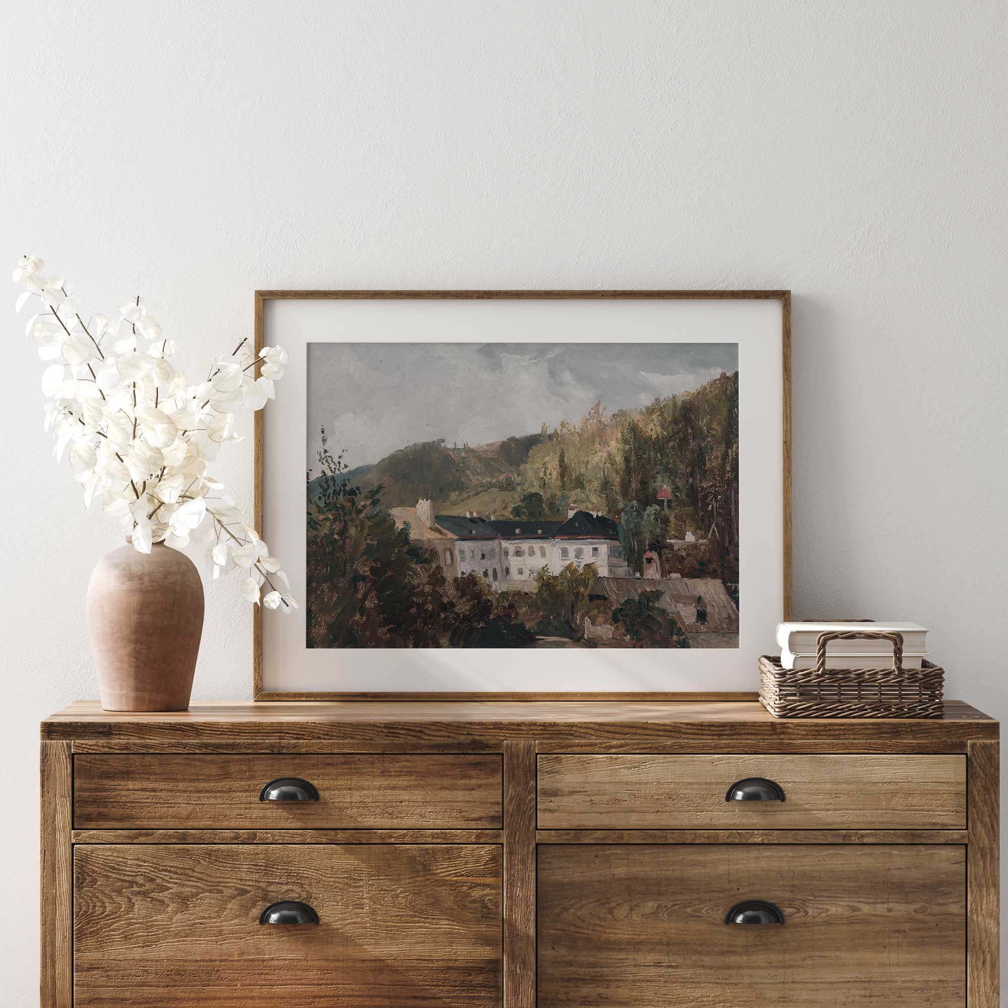 Wooden dresser with a framed Vintage Estate Print of European Manor in Woodland, vase, and basket on a white wall.