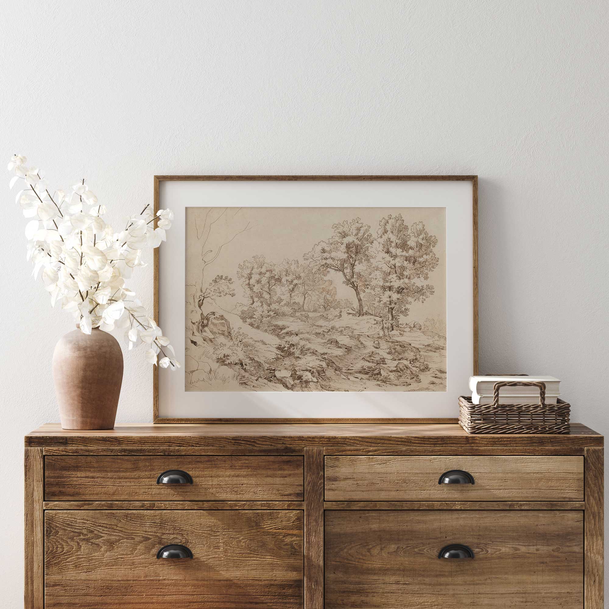 Wooden dresser with framed landscape drawing print, vase, and basket against a white wall