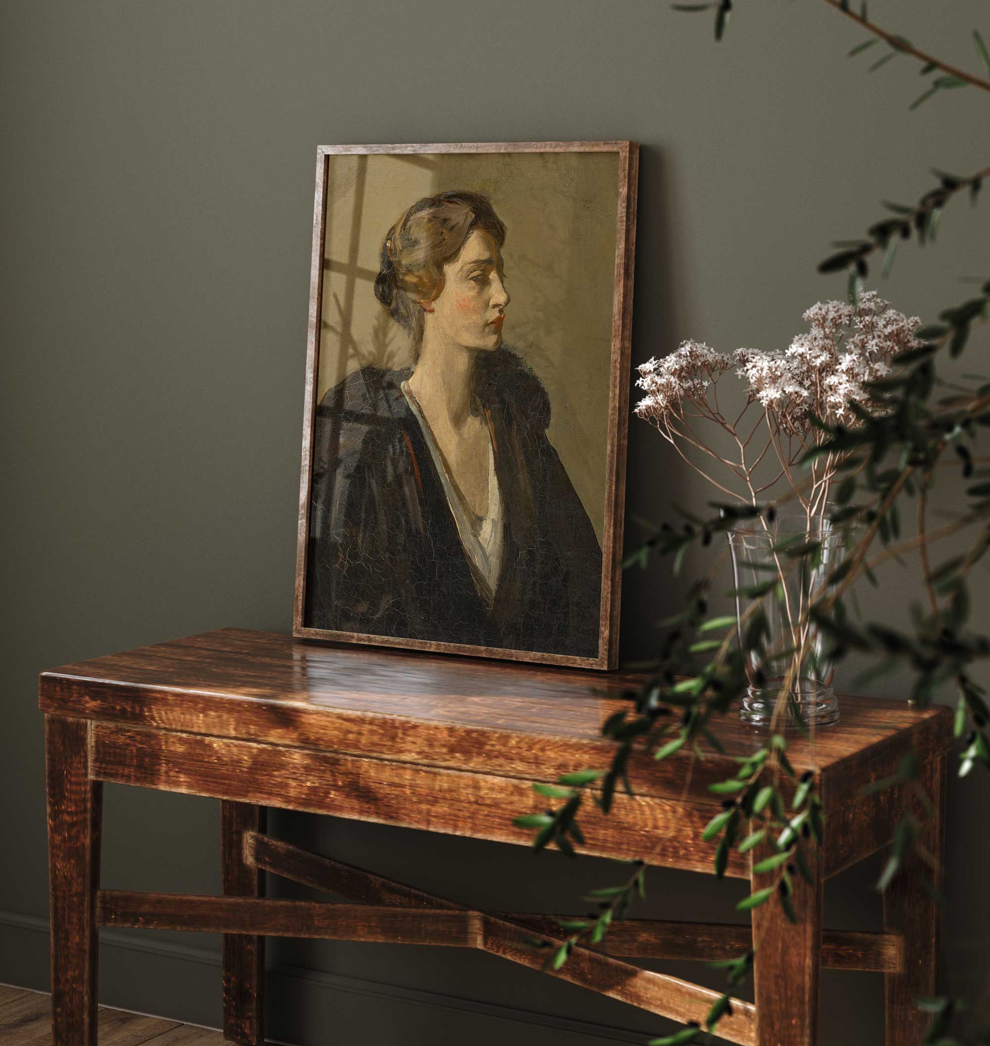 Framed portrait on a wooden table with a vase of flowers against a dark green wall.