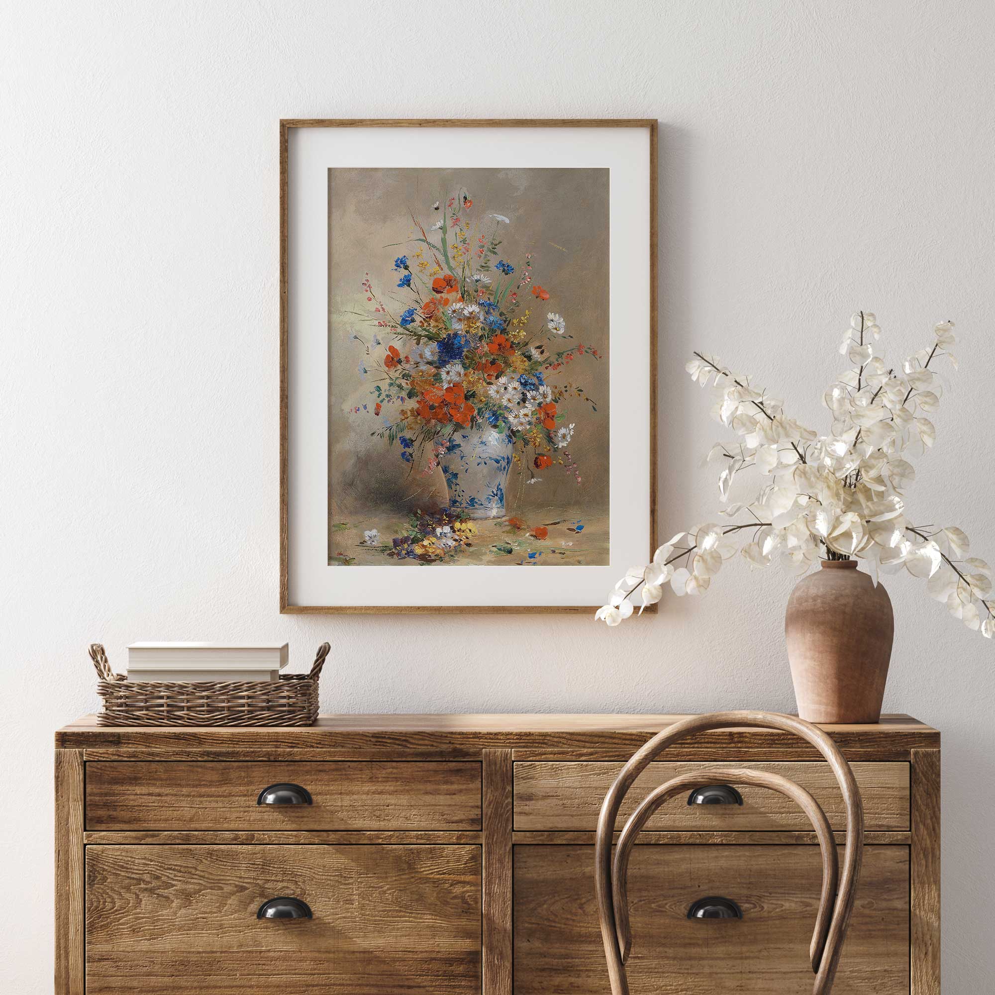 Wooden dresser with a framed floral painting above it, a vase with flowers to the right, and a basket on top.