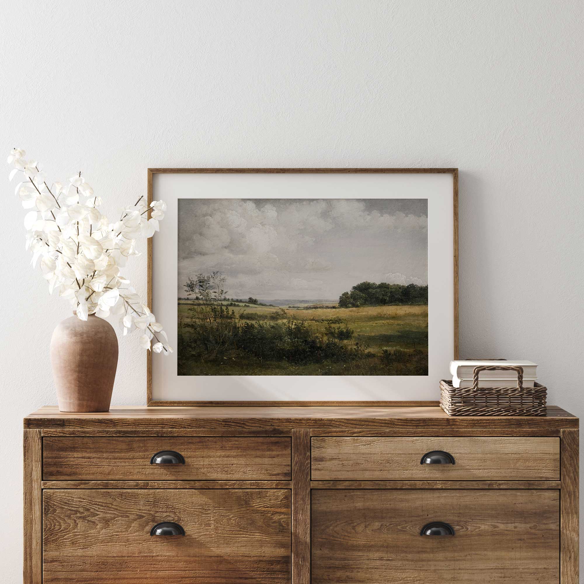 Wooden dresser with a Rustic Landscape Print of Golden Fields and Sunlit Clouds, vase, and basket on a white wall.