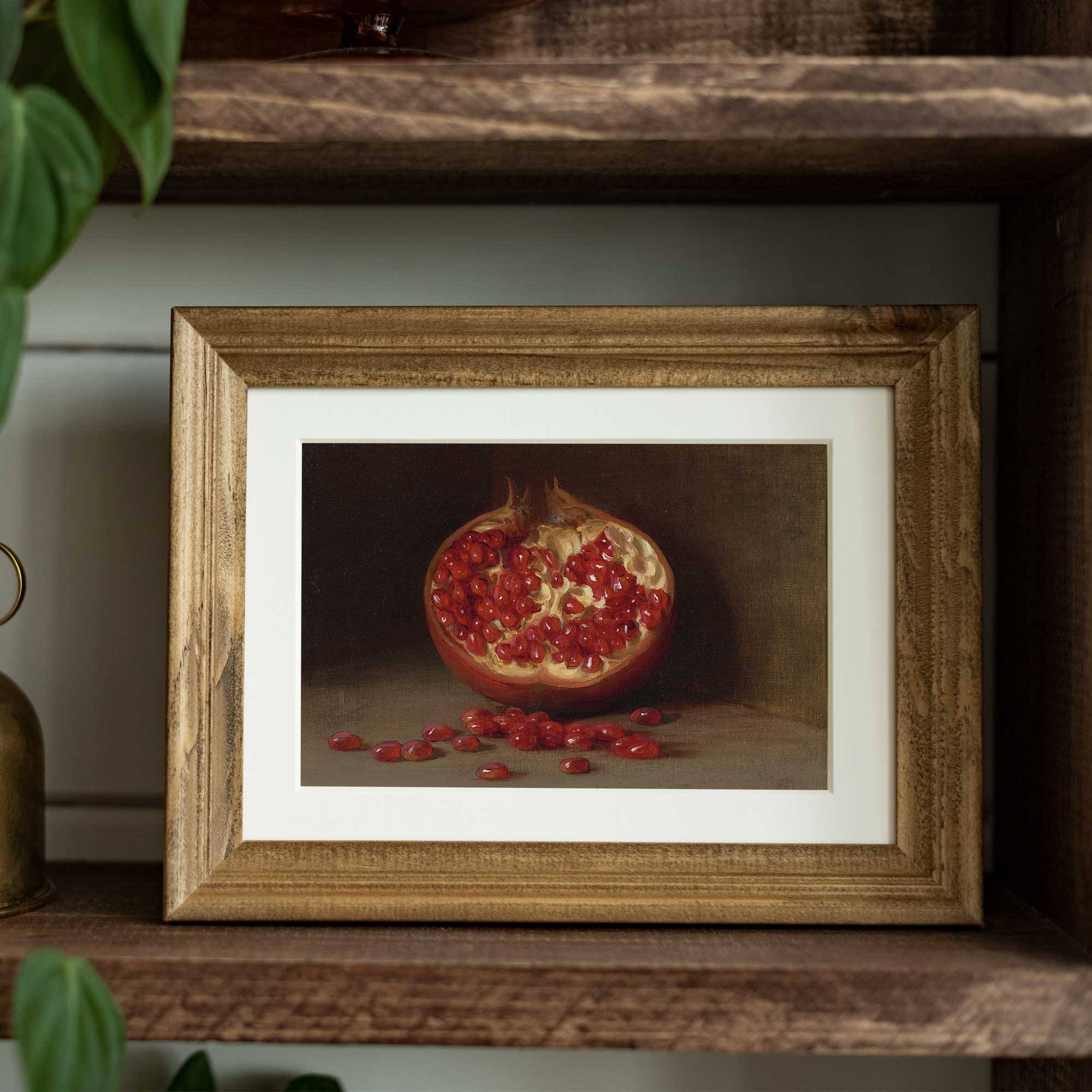 Framed still life painting of a pomegranate on a wooden shelf.