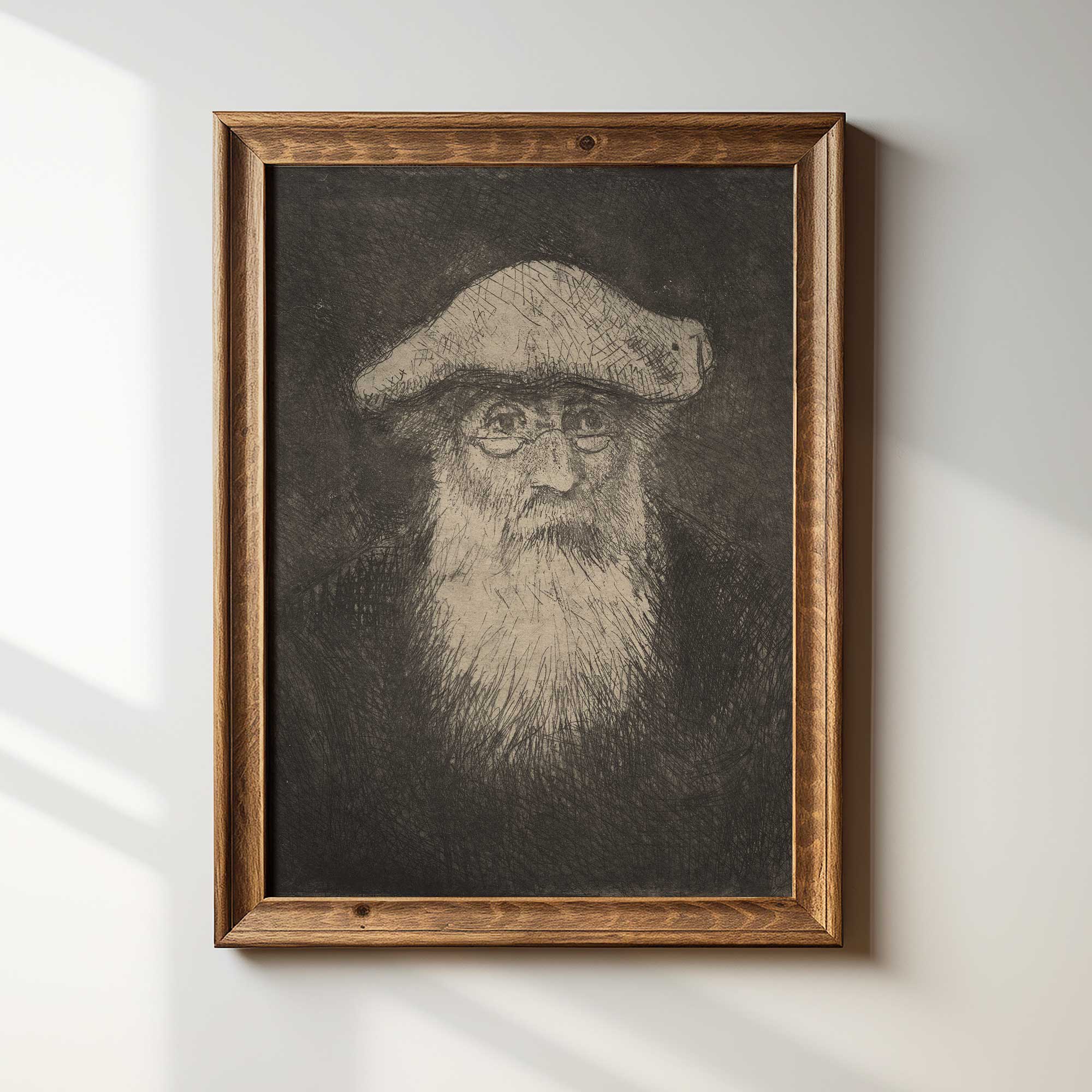 Framed black and white portrait of Camille Pissarro with a beard and hat on a light wall.