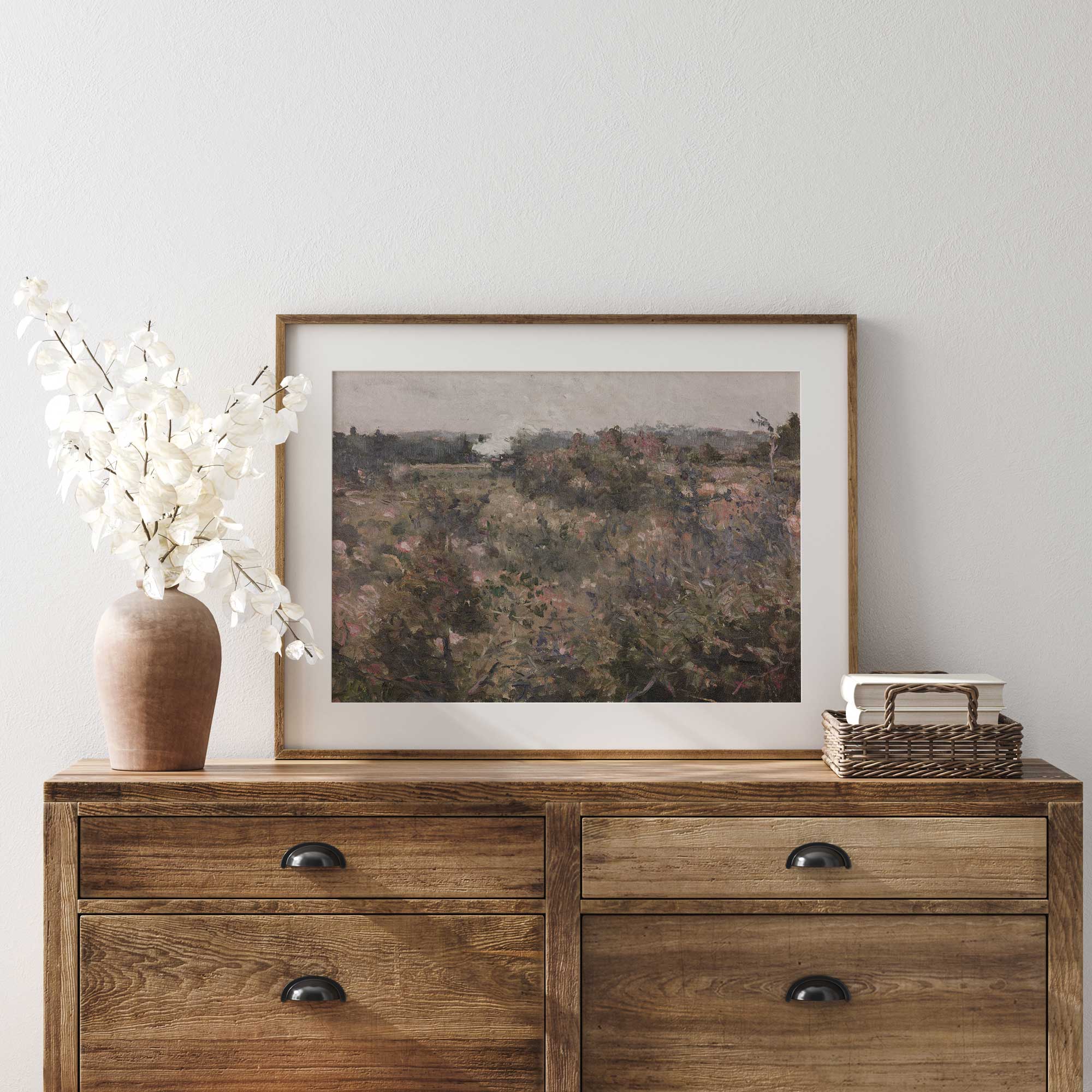 Wooden dresser with framed French Meadow Field Landscape Print , vase, and basket against a white wall