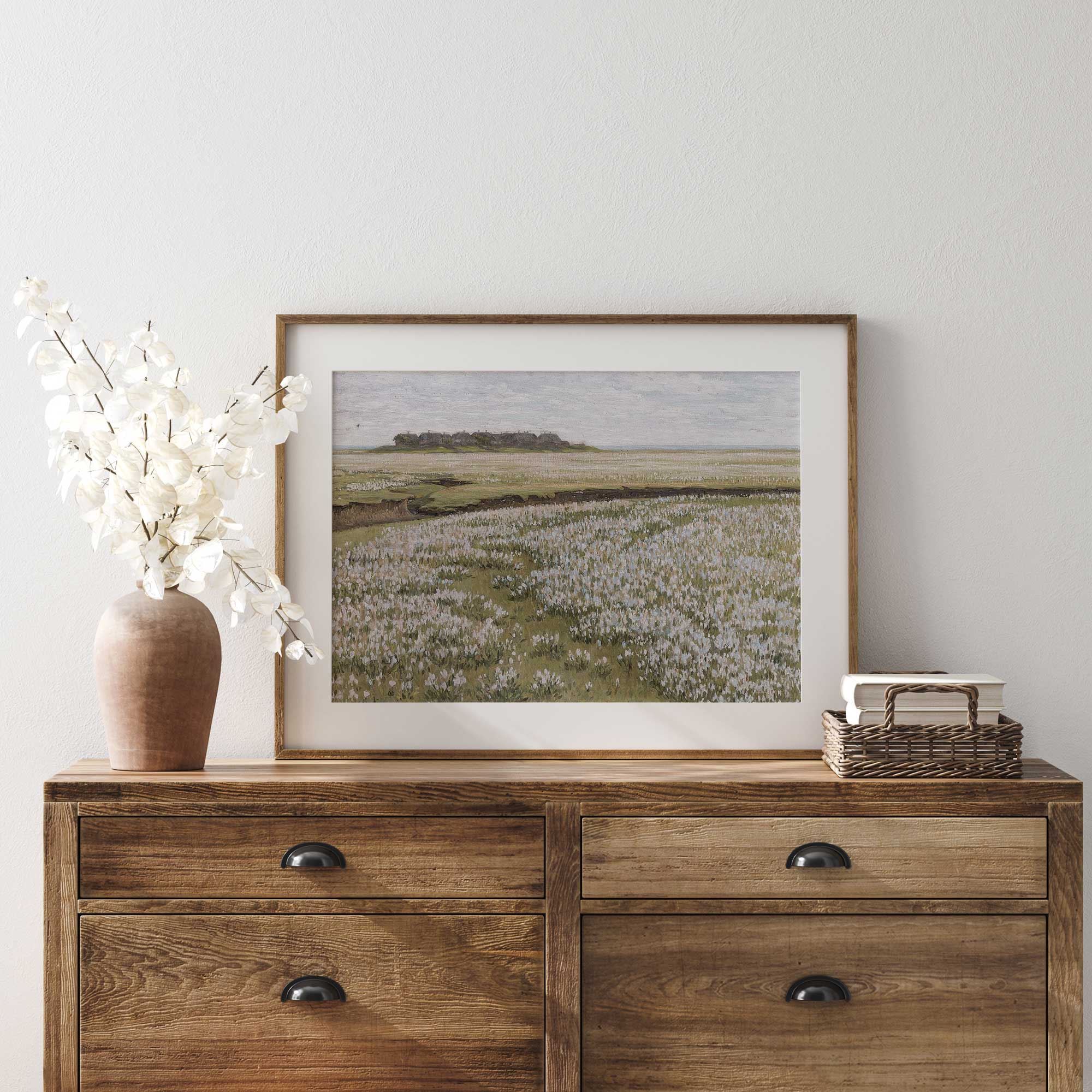 Wooden dresser with a framed Blooming Floral Meadow landscape print, vase, and basket on a white wall.