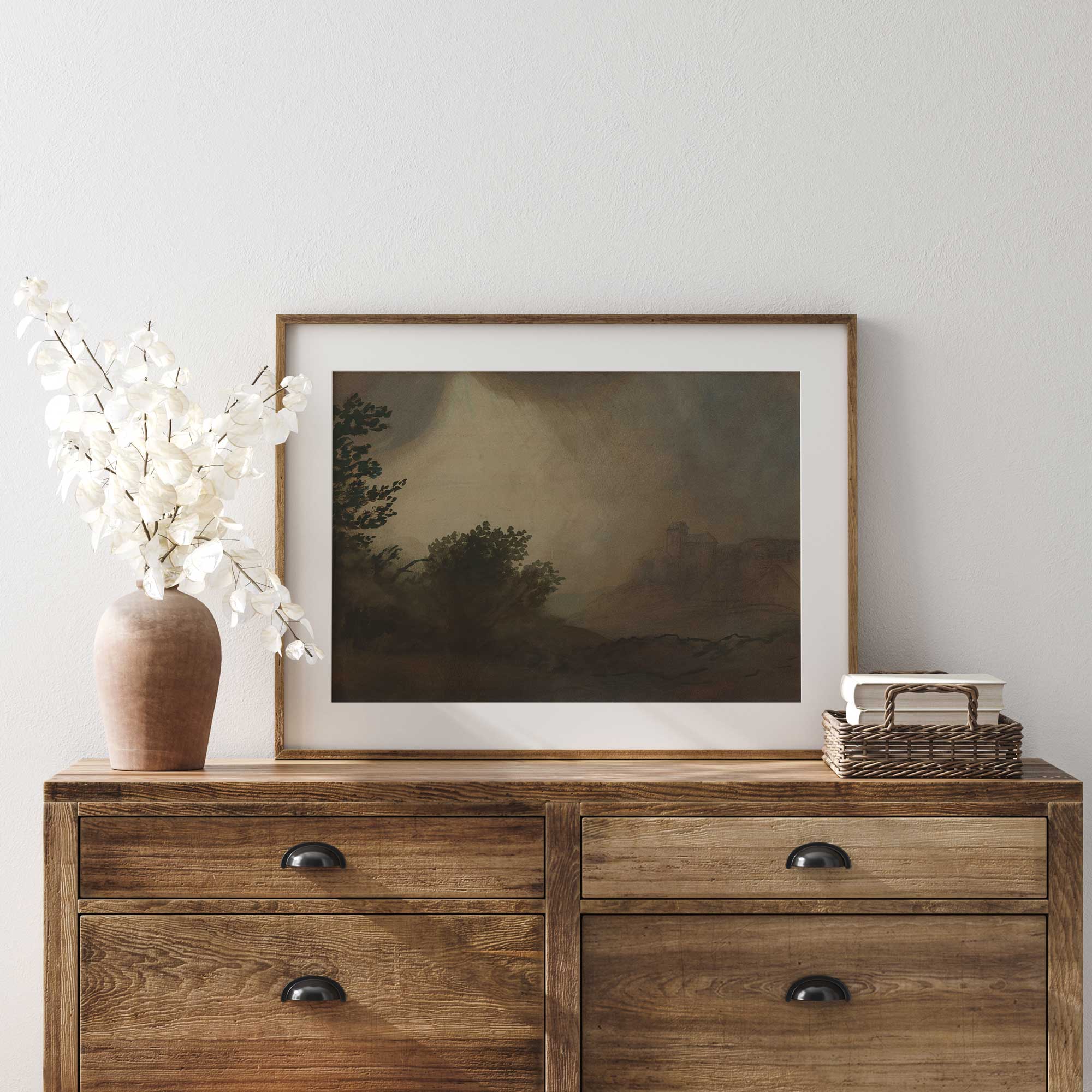 Wooden dresser with a framed Dark Vintage Landscape Print, vase, and books against a white wall.