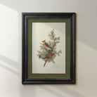 Framed artwork of two birds perched on a branch with leaves on a white wall.