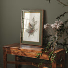 Framed artwork of a bird on a branch with greenery on a wooden table against a dark wall.