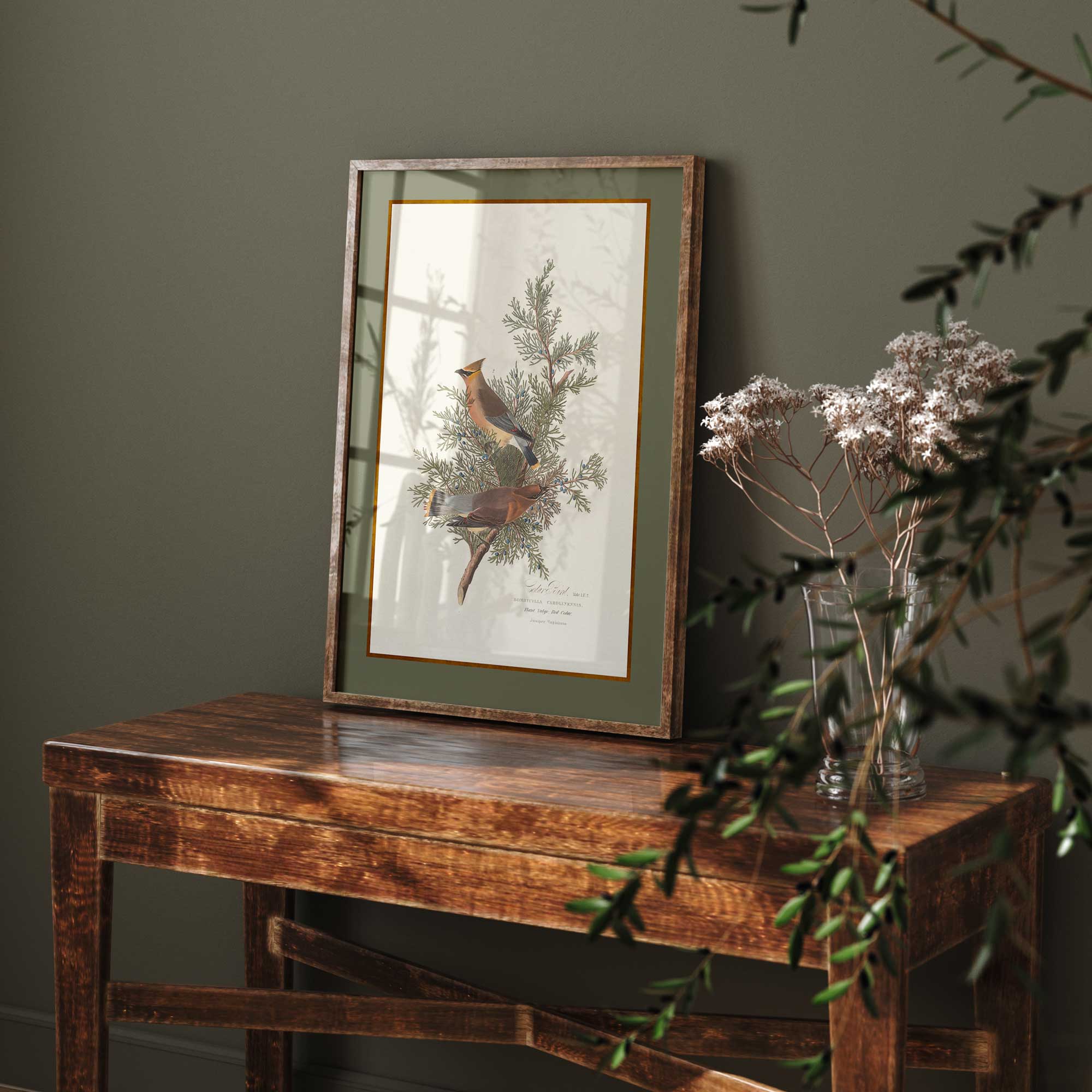 Framed artwork of a bird on a branch with greenery on a wooden table against a dark wall.