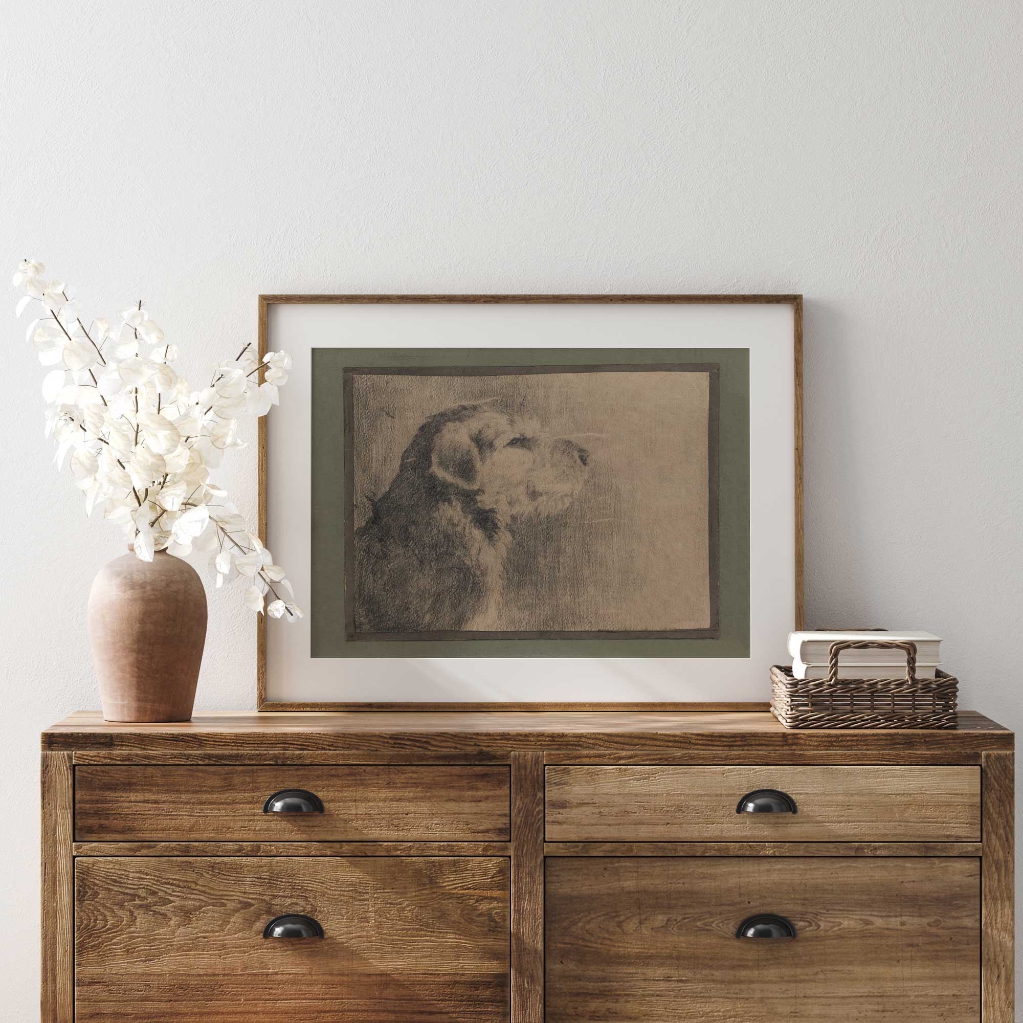 Wooden dresser with framed artwork of a dog, vase with flowers, and stack of books against a white wall.