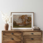 Wooden dresser with a framed autumn vintage landscape print,  vase, and decorative items against a white wall.