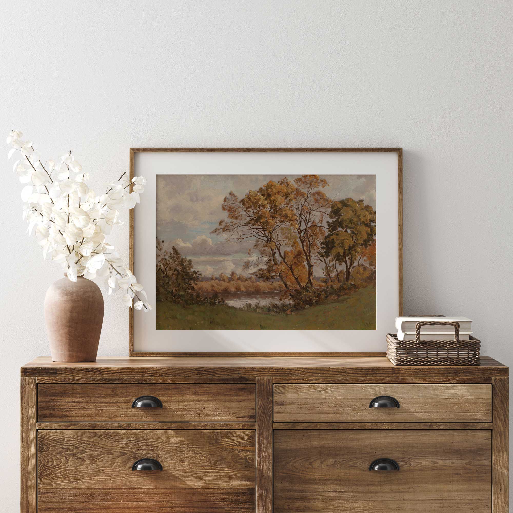 Wooden dresser with a framed autumn vintage landscape print,  vase, and decorative items against a white wall.