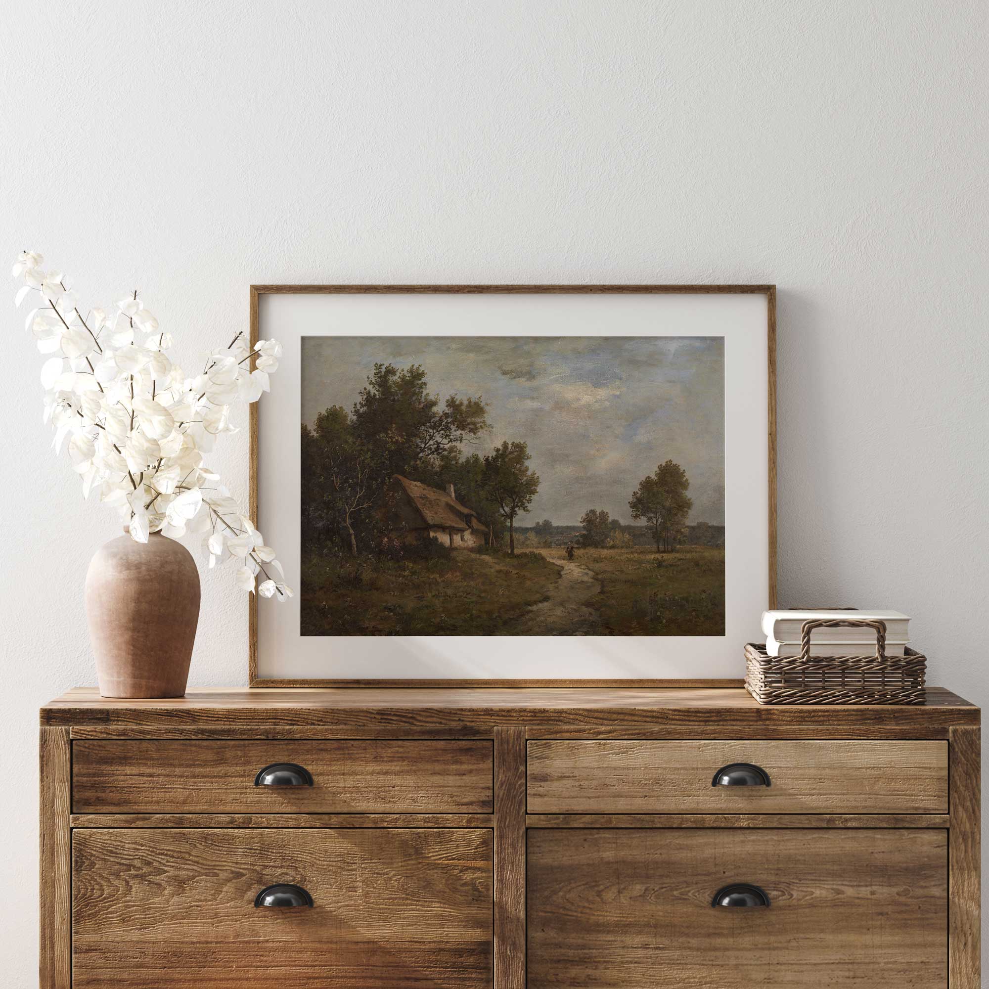 Wooden dresser with a framed Rustic country landscape print, vase, and books against a white wall.

