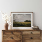 Wooden dresser with a Rustic Landscape Print of Golden Fields and Sunlit Clouds, vase, and basket on a white wall.