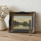 Framed French landscape Wall Art on a wooden surface with a vase of white flowers.