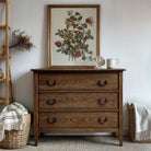 Wooden dresser with floral painting above, wicker baskets on either side, and a neutral color scheme.